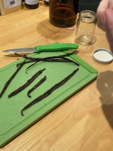 Vanille Schoten ganz auf einem grünen Schneidebrett, mit Messer, Glas und Öl im HIntergrund